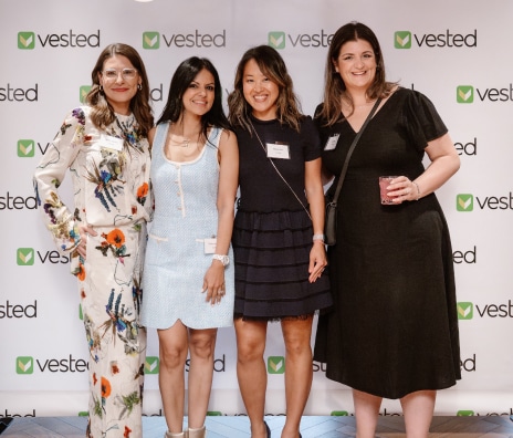 Four women smiling together in front of a Vested branded step-and-repeat backdrop at a company event