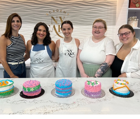 Five women posing with decorated cakes at a team cake decorating class at NY Cake Academy
